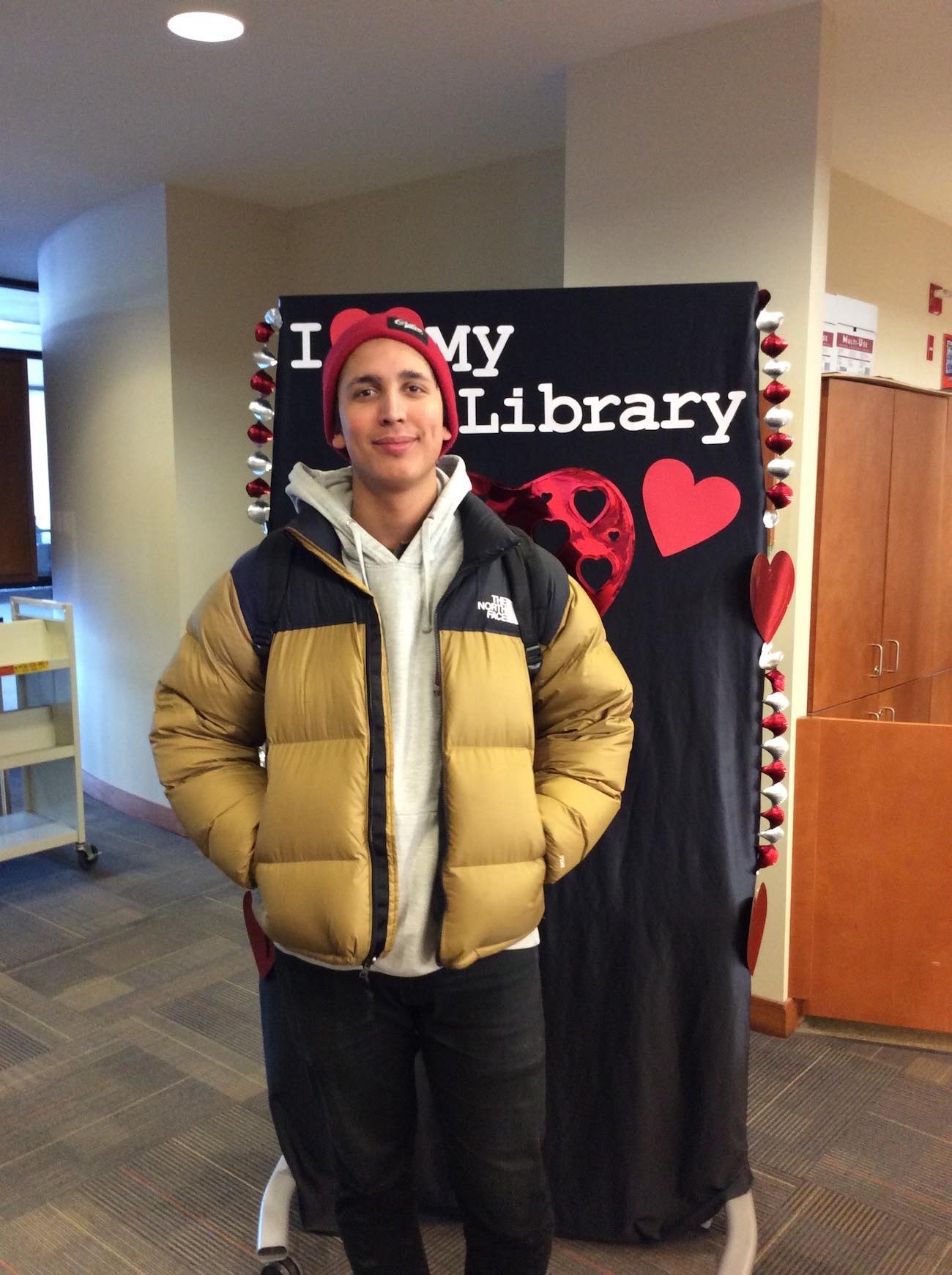 A smiling student in a red toque and yellow puffy jacket stands in front of the I love my library backdrop with his hands in his pockets.